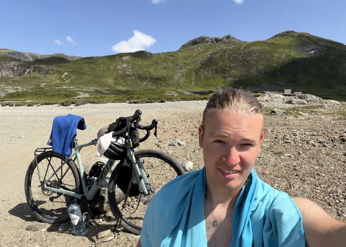 Selfie mit Handtuch um den Hals, dahinter das bepackte Rad und grüne Berge